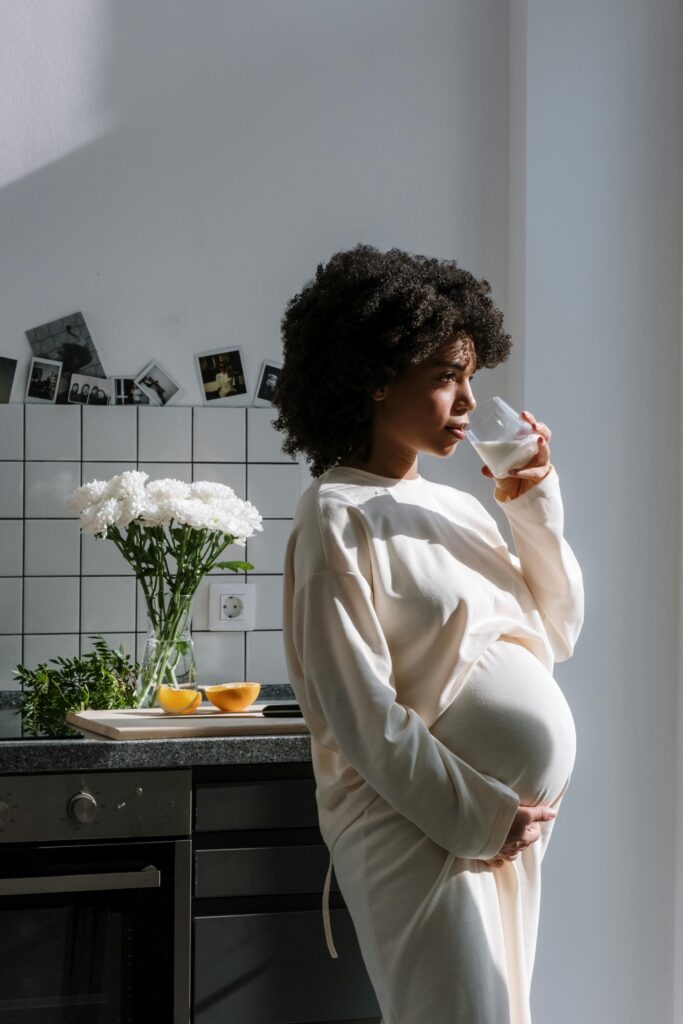 Pregnant woman drinking milk in a sunlit kitchen, surrounded by flowers and fruit.